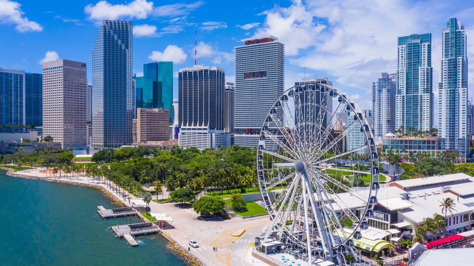 A coastal cityscape with a prominent Ferris wheel, high-rise buildings, and a marina.