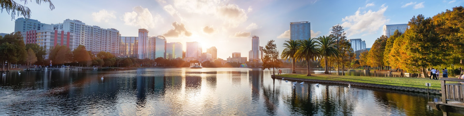 Paisagem urbana com um corpo d'água refletindo edifícios, um parque com árvores e um céu claro.
