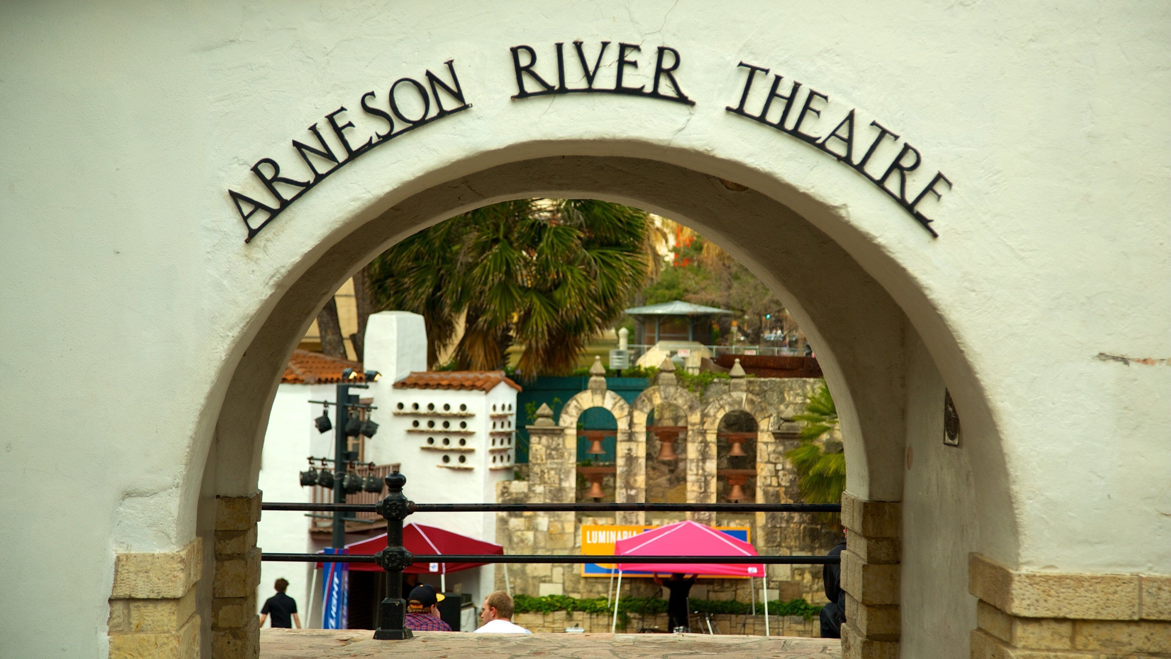 An archway with the inscription "ARNESON RIVER THEATRE" above a view of a street scene with buildings and a palm tree.
