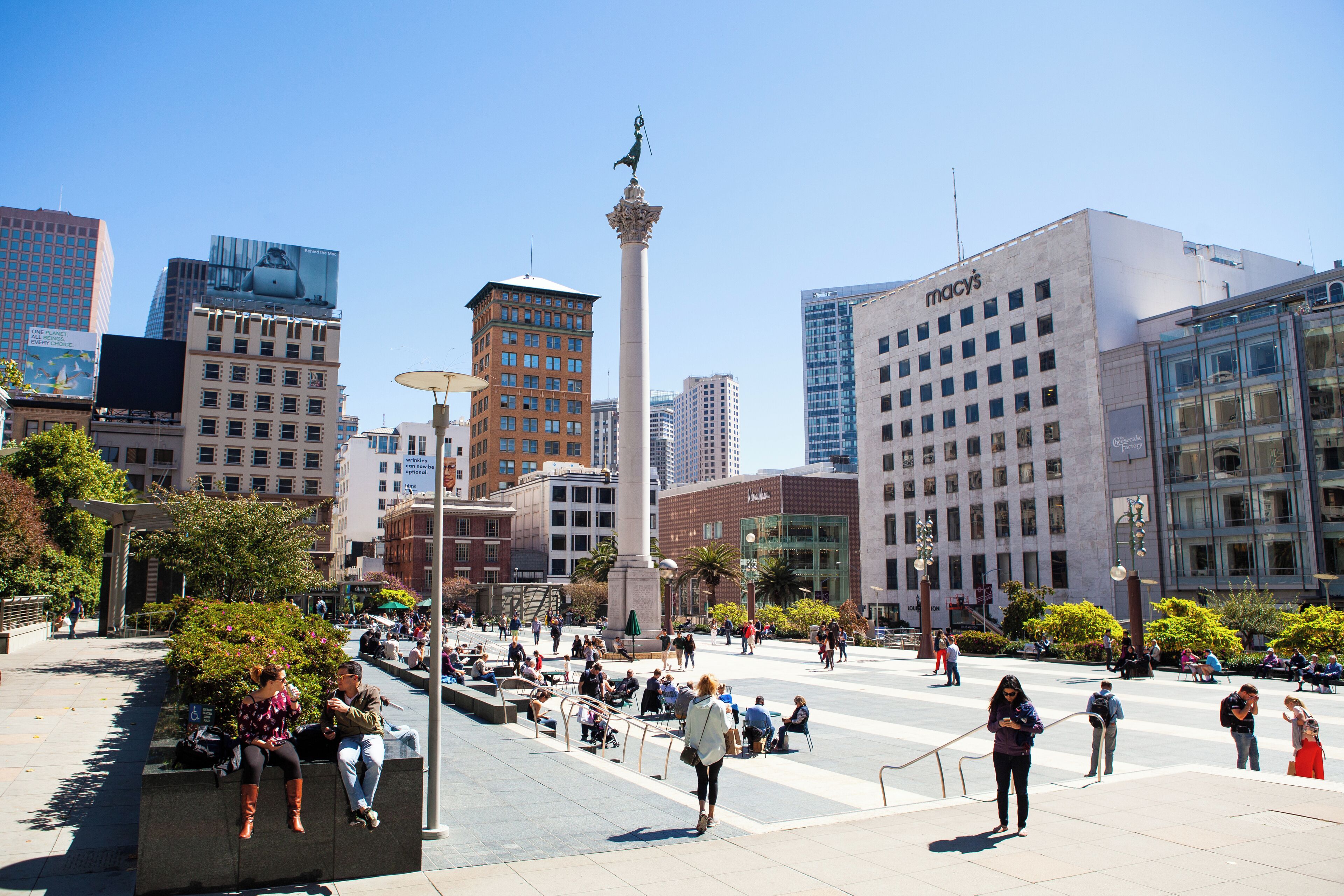 Una plaza urbana con un monumento imponente, edificios modernos y gente sentada y caminando.