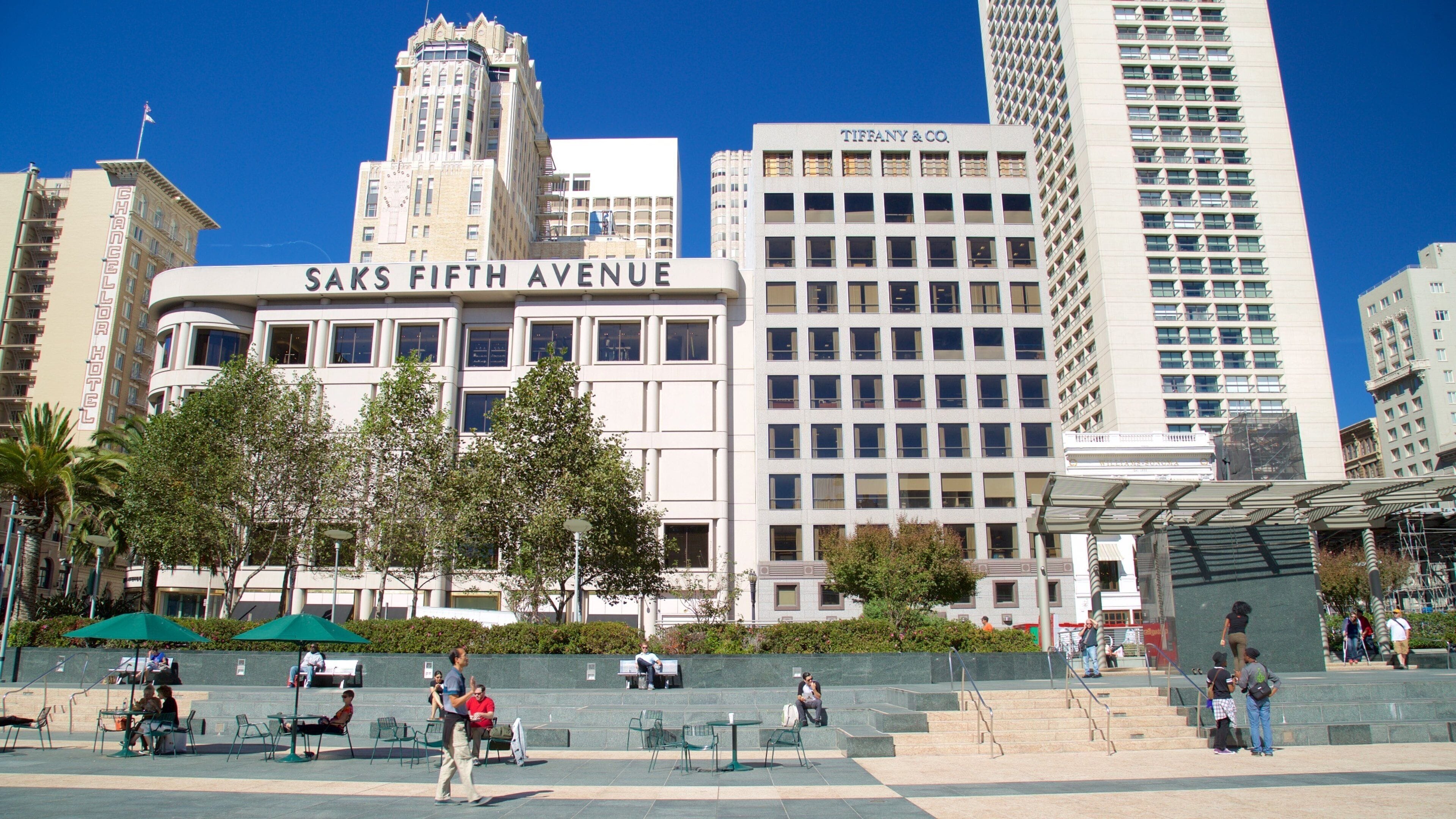 Saks Fifth Avenue, un imposant bâtiment blanc aux grandes fenêtres, est entouré d’autres immeubles de grande hauteur et de palmiers.