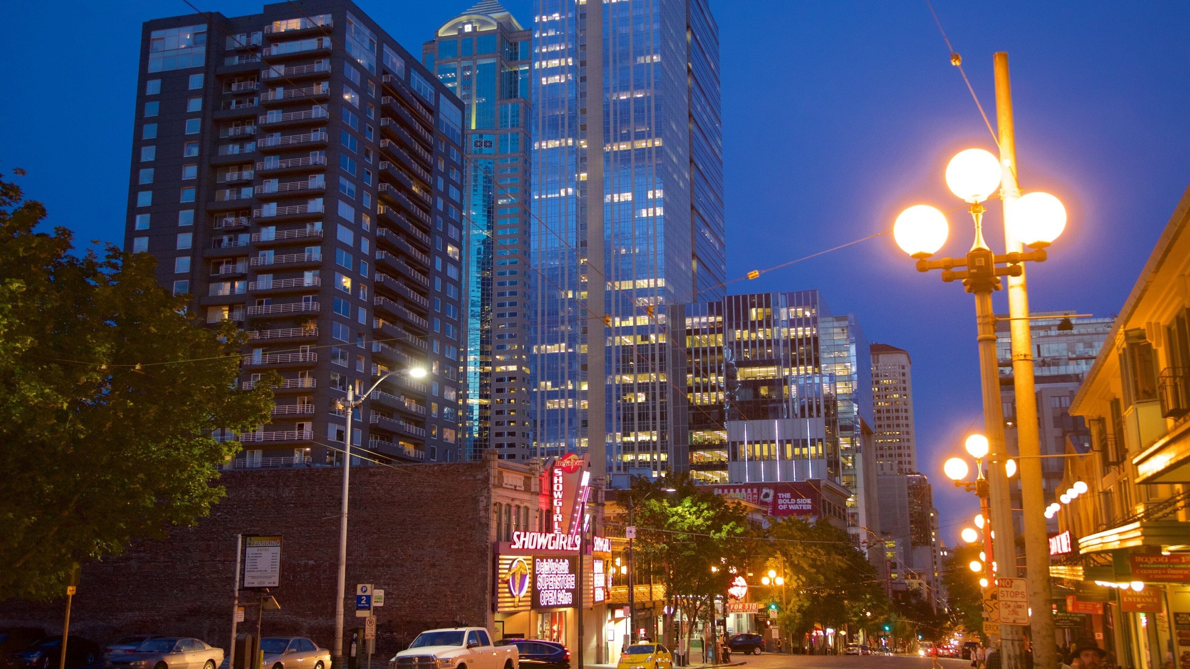 A cityscape at dusk with illuminated buildings, street lamps, and a neon sign for 'Showgirls'.