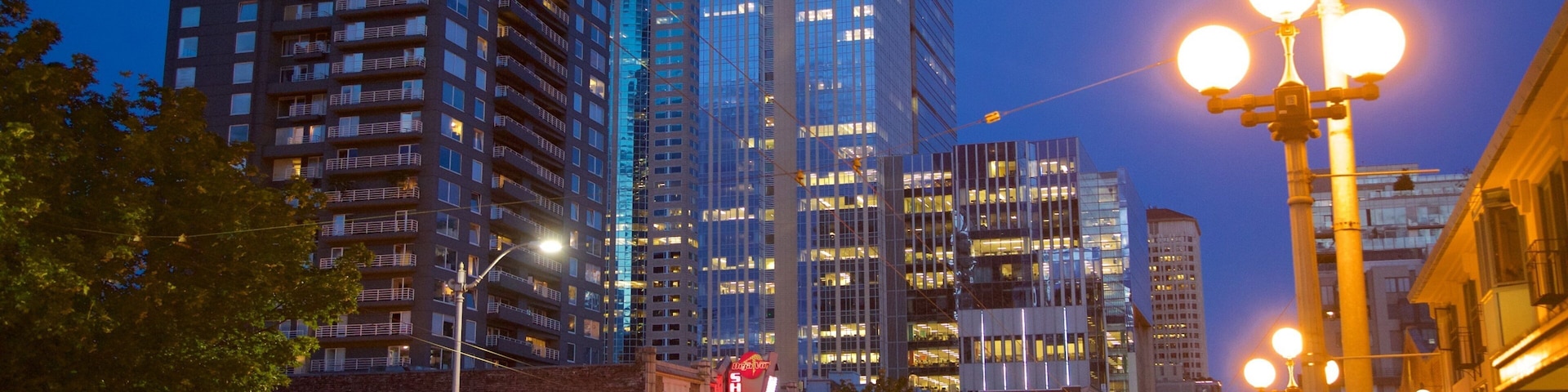 Paisagem urbana ao entardecer com edifícios iluminados, postes de luz e um letreiro de neon com a inscrição 'Showgirls'.