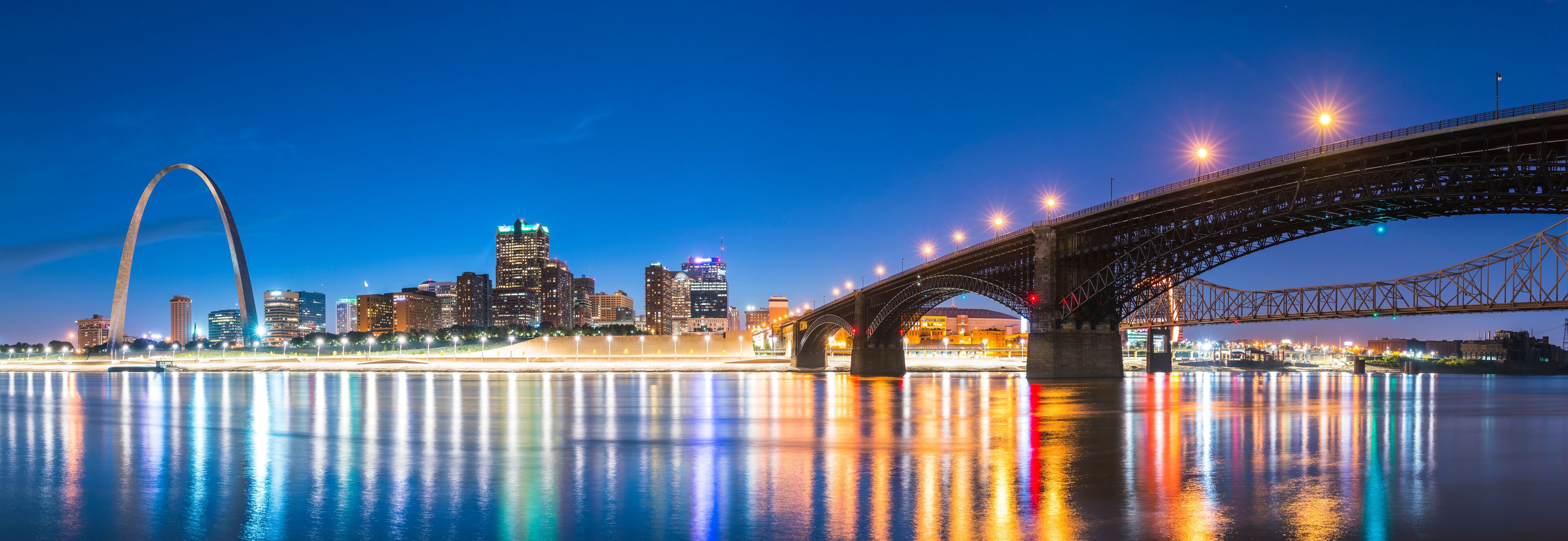 A city skyline with a prominent arch bridge over a river reflecting city lights.