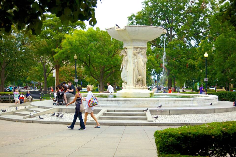 A public park with a central fountain, benches, and people walking around.