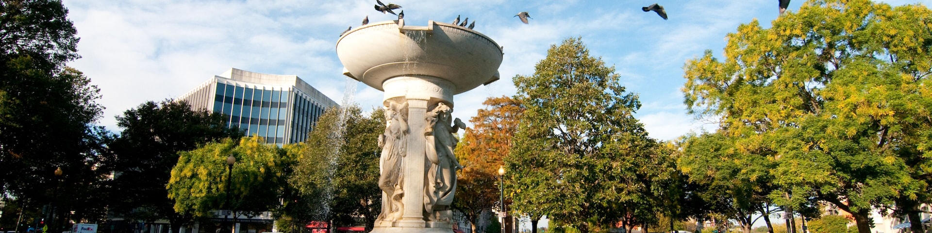 A fountain with a statue base, surrounded by trees and a modern building in the background.