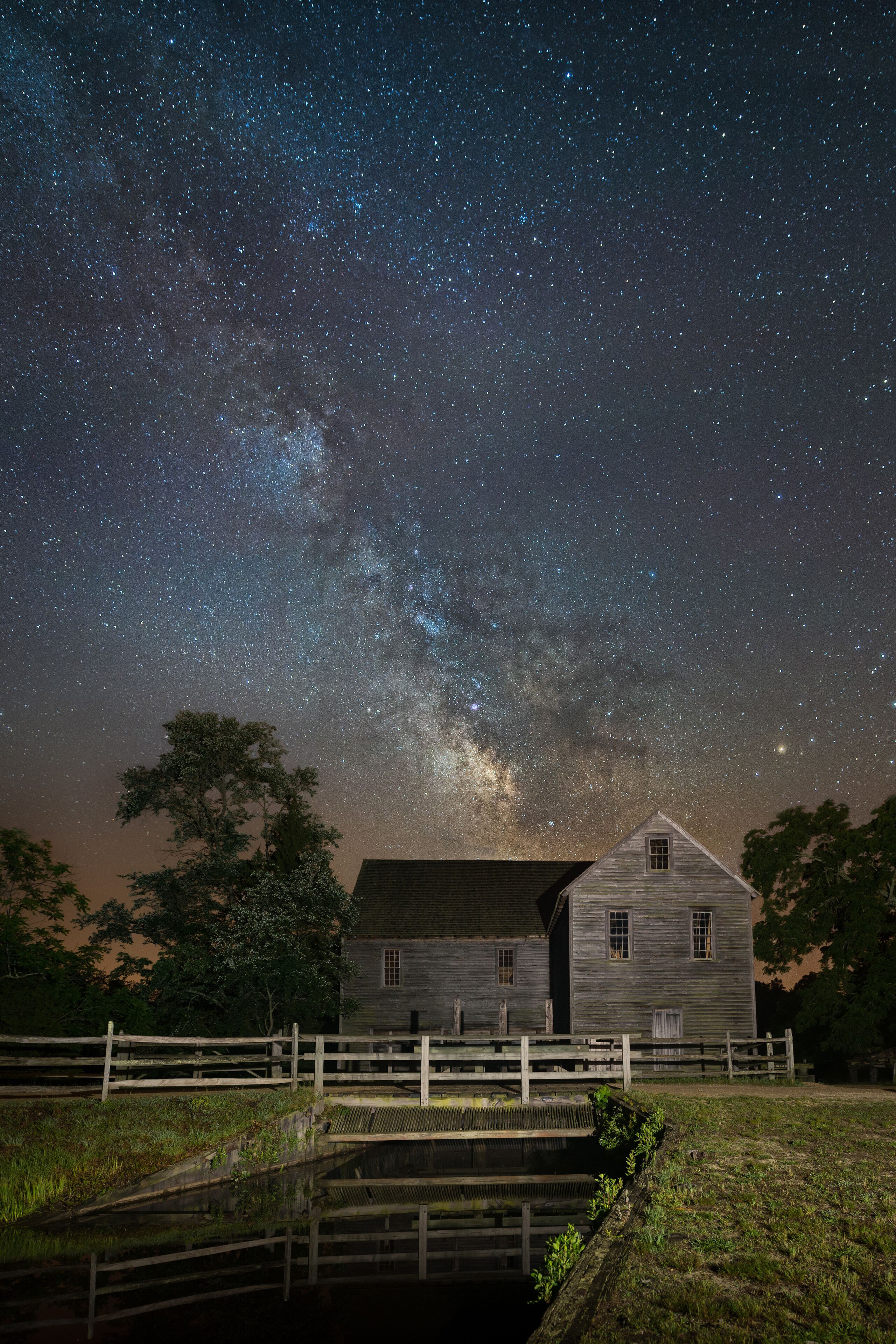A rustic wooden house with a wooden fence, a small pond, and a starry night sky.
