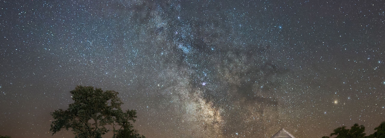 Una casa de madera rústica con valla de madera, un pequeño estanque y un cielo nocturno estrellado.