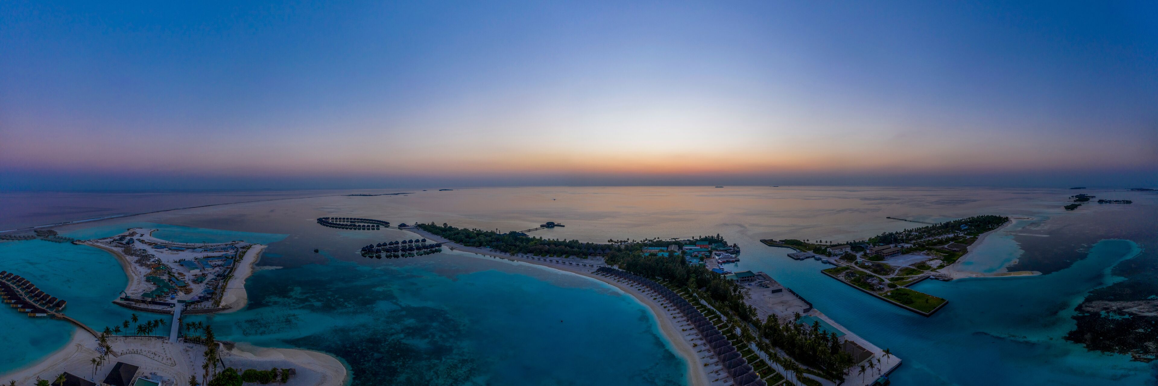 An aerial view of a coastal resort with clear turquoise waters, a crescent-shaped beach, and a circular lagoon with overwater bungalows.