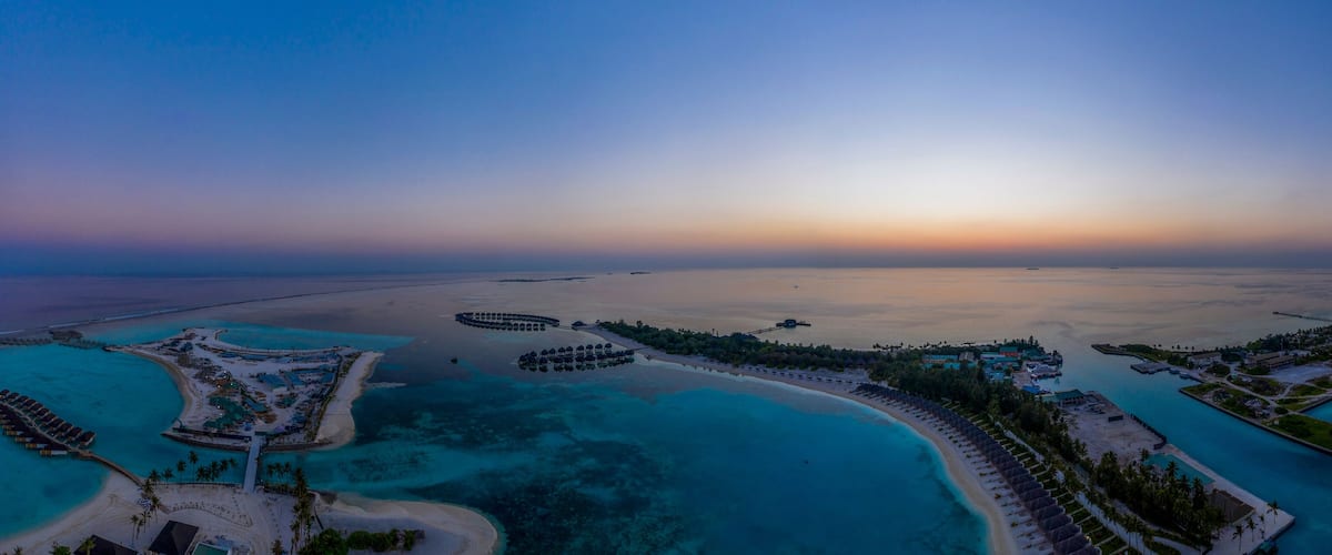 An aerial view of a coastal resort with clear turquoise waters, a crescent-shaped beach, and a circular lagoon with overwater bungalows.