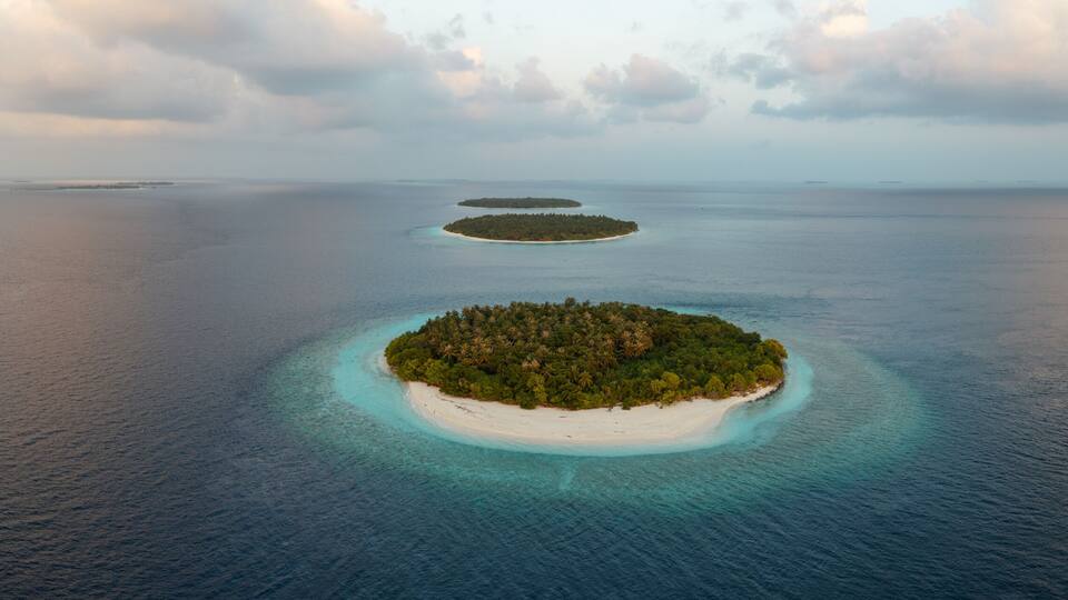 An aerial view of a tropical island surrounded by clear turquoise waters, with a dense forest covering the island and smaller islets in the distance.