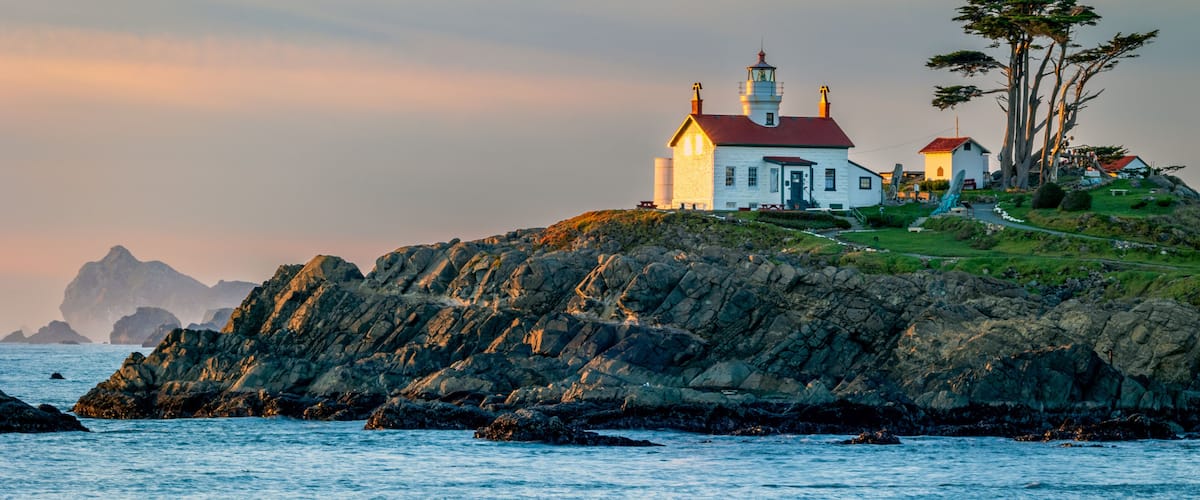Un faro costero, una costa rocosa y montañas a lo lejos.