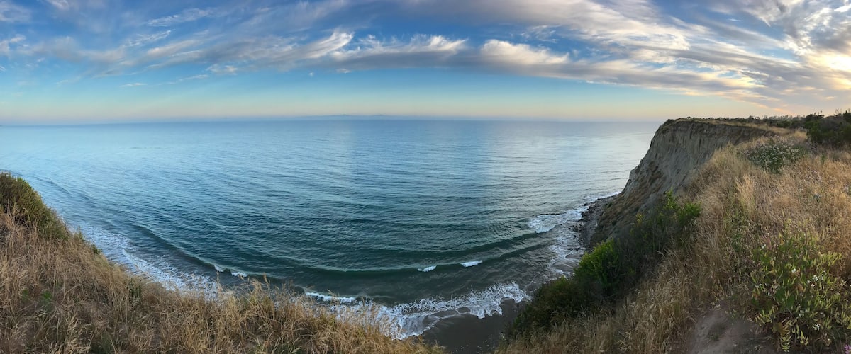 A coastal landscape with a cliff overlooking the ocean, waves crashing against the shore, and a clear sky.
