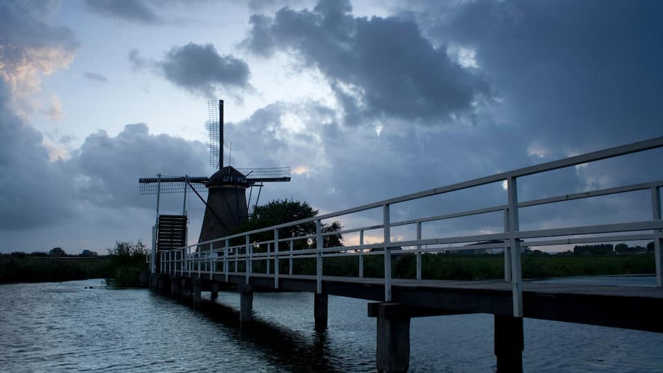 A windmill on a bridge over water, with a cloudy sky.
