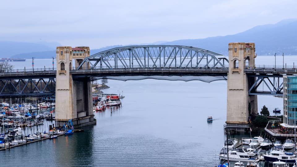 A large steel bridge spanning a calm river with boats docked at the marina.