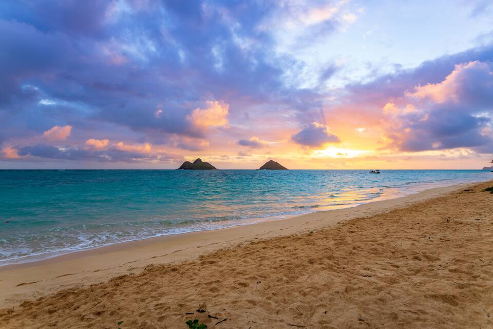A beach with clear turquoise water, a sandy shore, and two small islands in the distance during sunset.