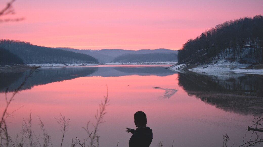 Adventurous woman pointing, standing in front of a pink sunrise on the horizon of Tygart Lake, West Virginia