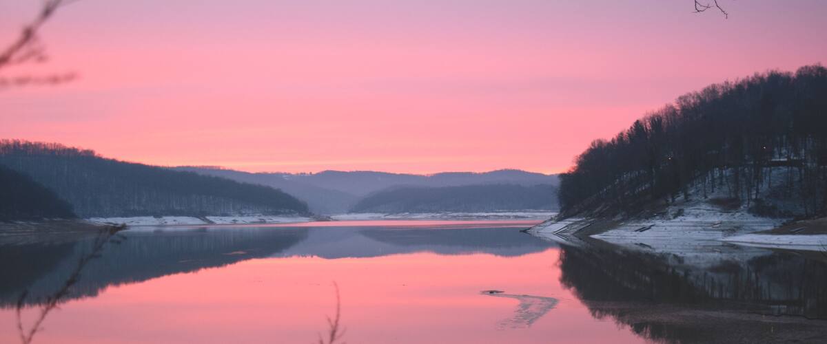 A person standing by a calm lake with a pink sky, surrounded by snow-covered hills and trees.