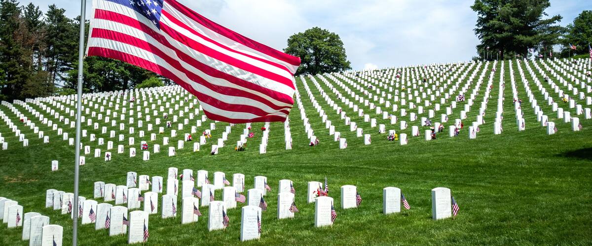 A cemetery with rows of white headstones and an American flag waving in the wind.