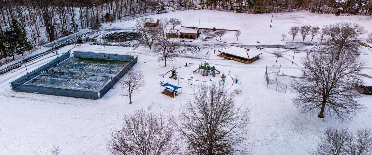 A snow-covered playground with swings and slides, surrounded by trees and buildings.