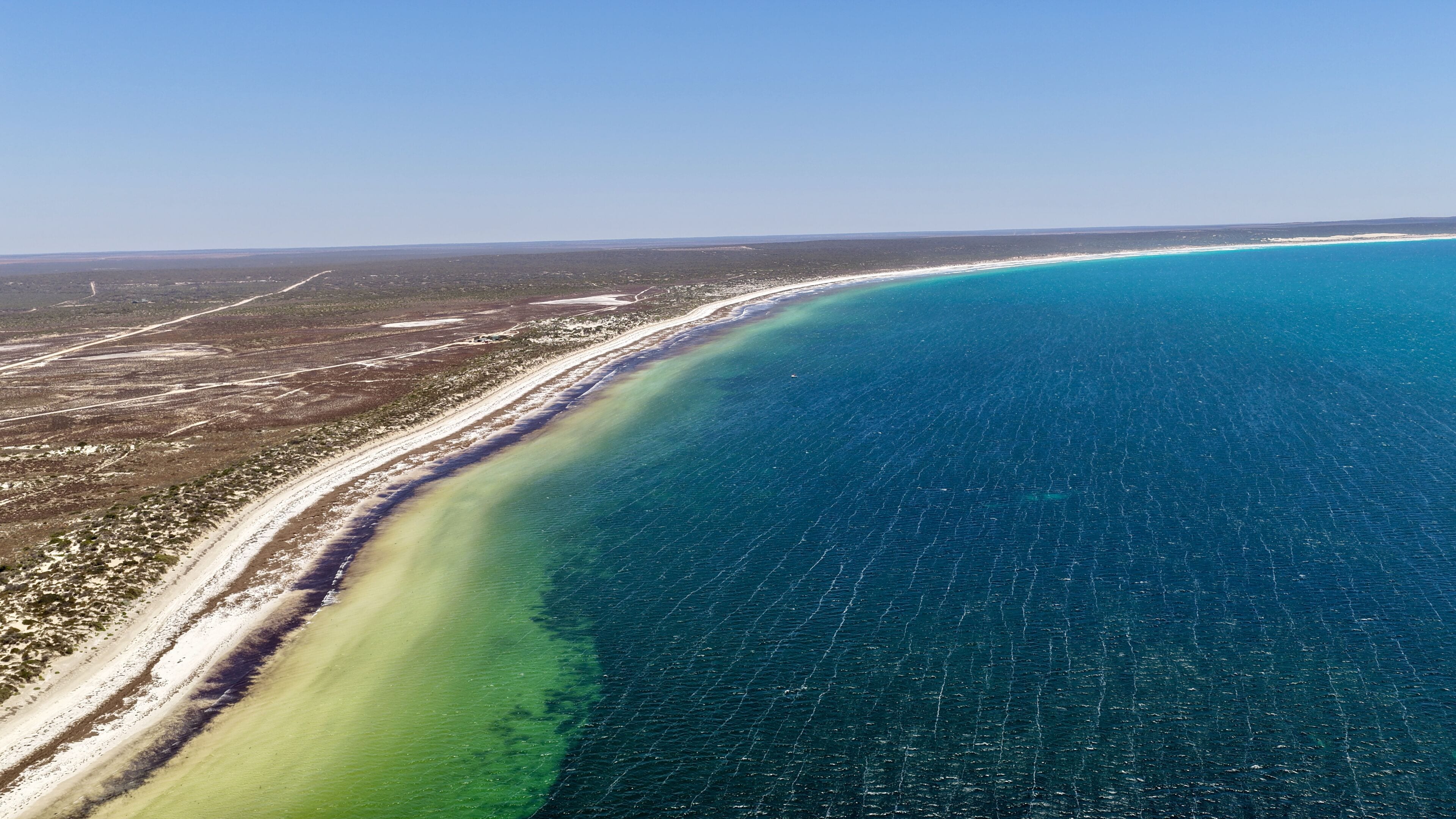 A coastal landscape with a long sandy beach, clear blue waters, and a distant horizon.