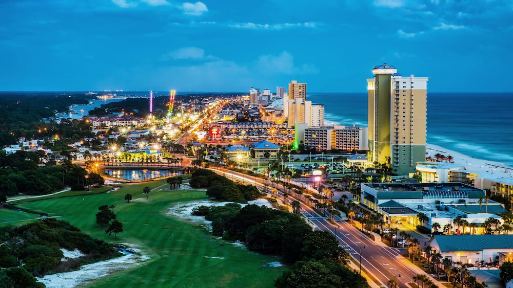 A coastal cityscape at dusk with illuminated streets, a beach, and a golf course.