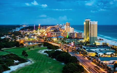 A coastal cityscape at dusk with illuminated streets, a beach, and a golf course.