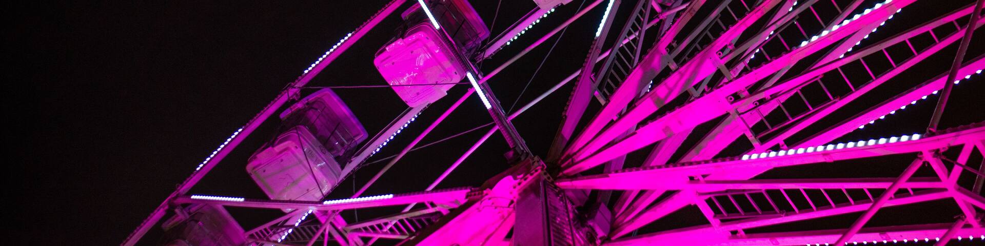 Close up of a bright pink ferris wheel lit up against a dark sky during a festival in a city