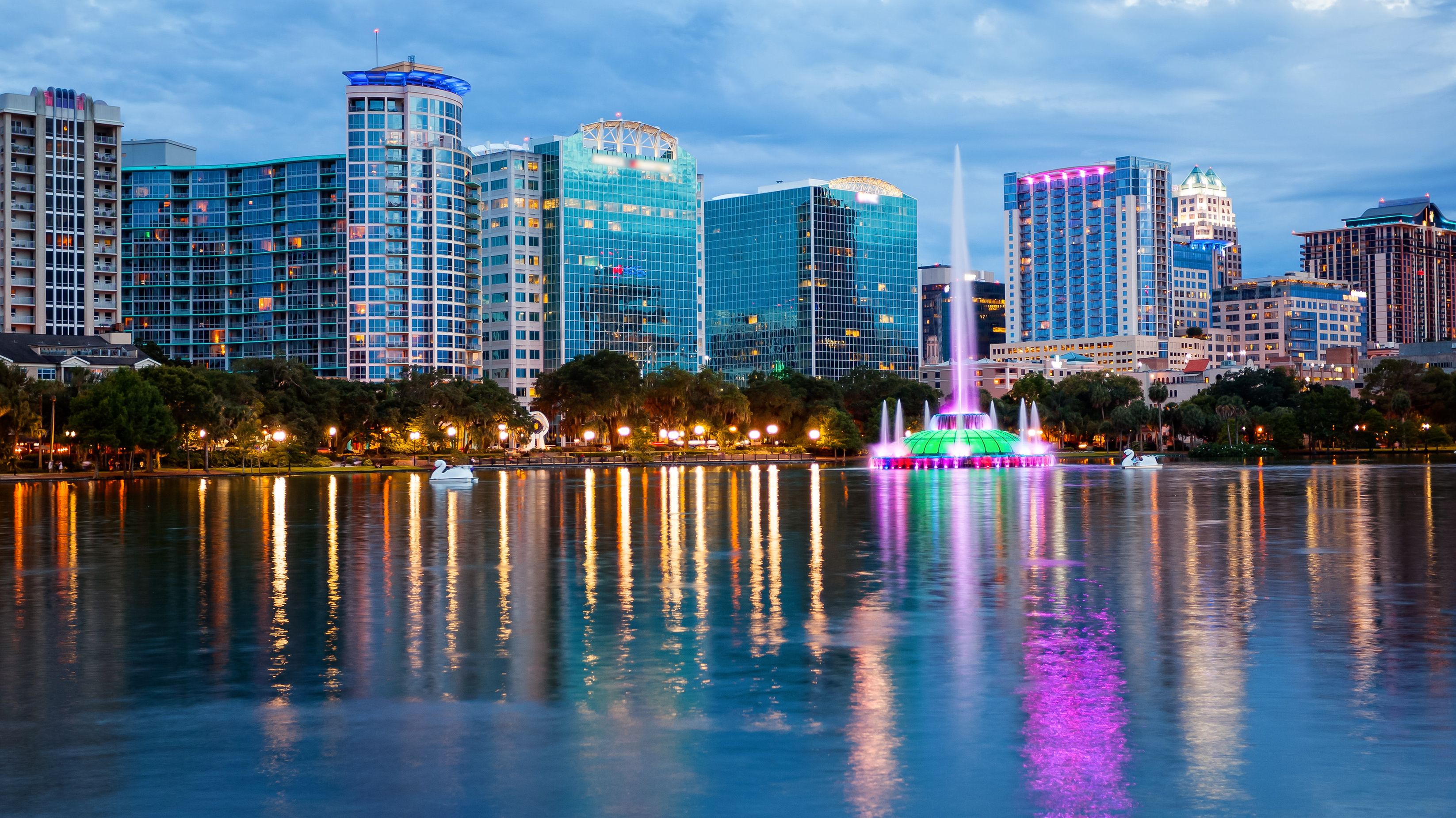 A city skyline with modern buildings, a body of water reflecting lights, and a fountain with colorful lights.