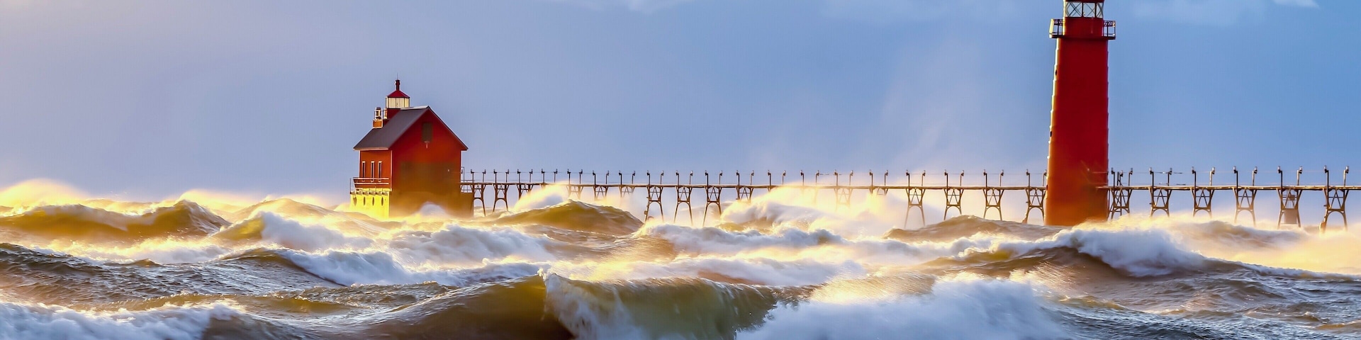 A red lighthouse and a pier extending into rough seas during sunset.