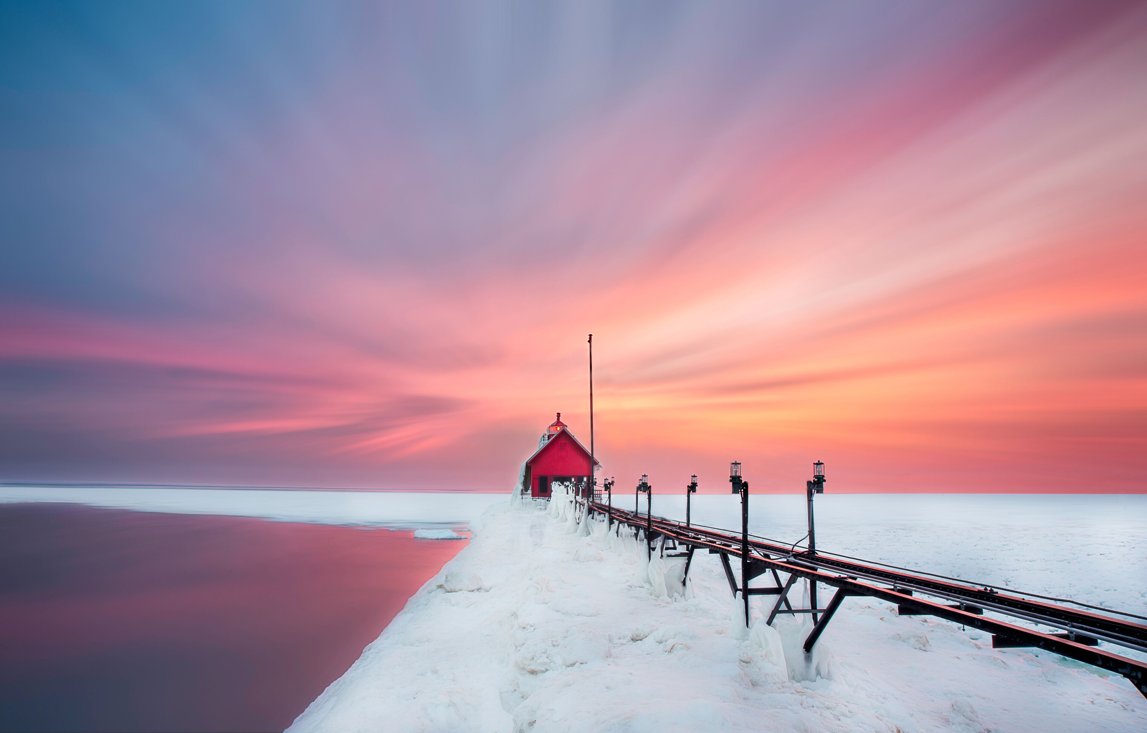 A red lighthouse on a snowy pier extending into a calm body of water, with a colorful sunset in the background.