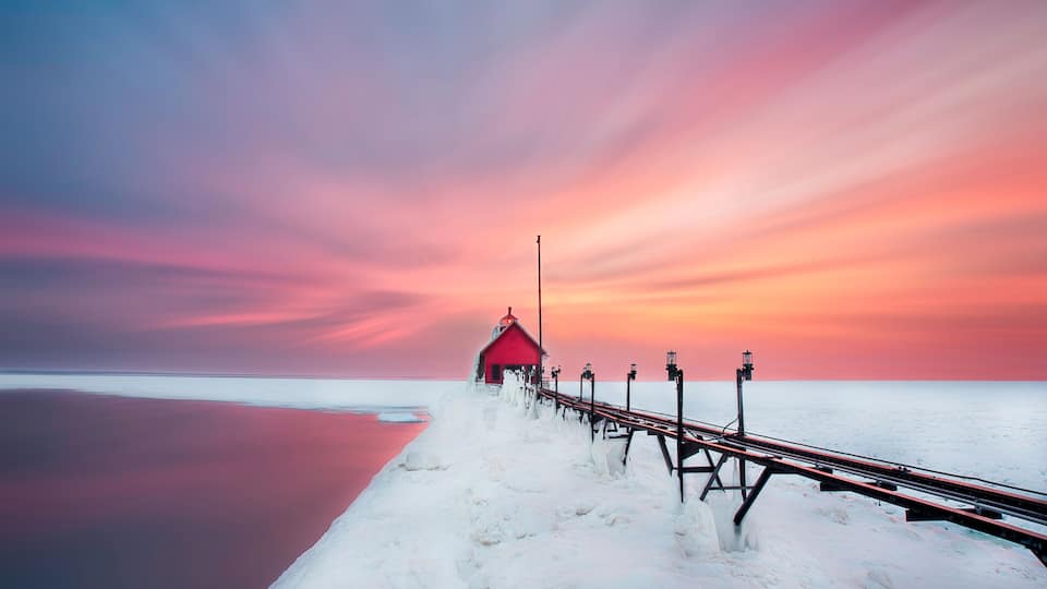 A red lighthouse on a snowy pier extending into a calm body of water, with a colorful sunset in the background.
