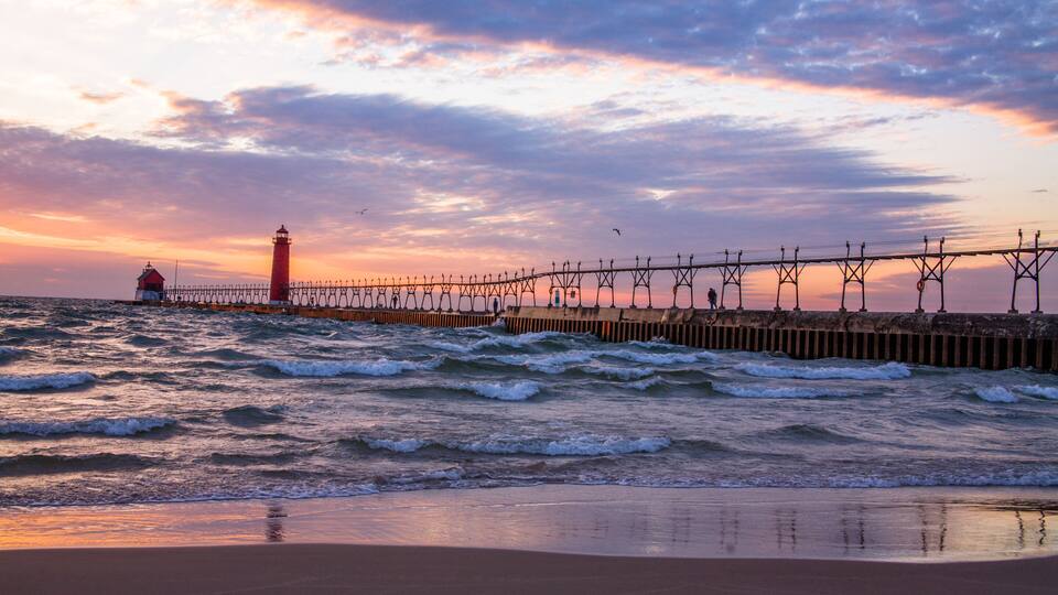 A coastal sunset with waves, a pier, and a lighthouse.