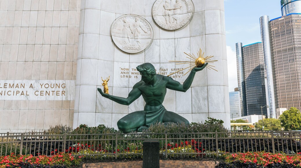 A statue of a muscular figure holding a radiant orb, set against a backdrop of a city wall with historical plaques and a modern skyscraper.