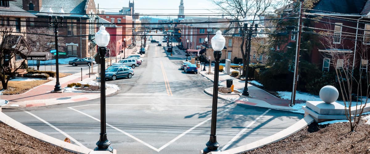 A town street with cars parked on both sides, a church steeple in the distance, and a roundabout with street lamps.