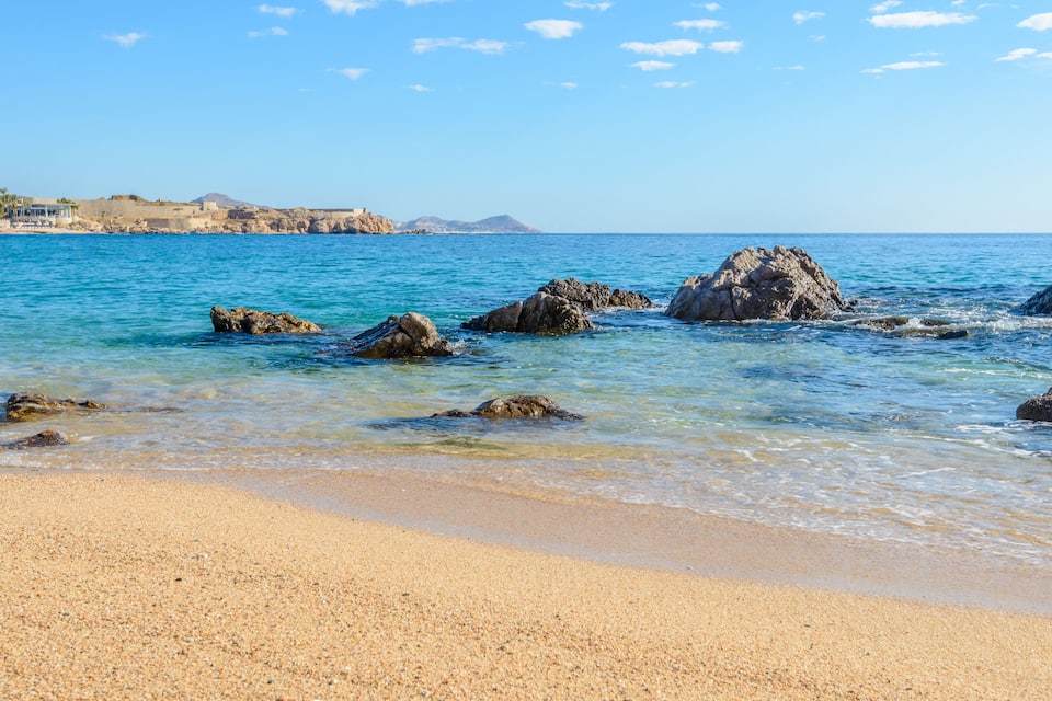 Una playa con agua turquesa cristalina, afloramientos rocosos y una cadena montañosa distante.