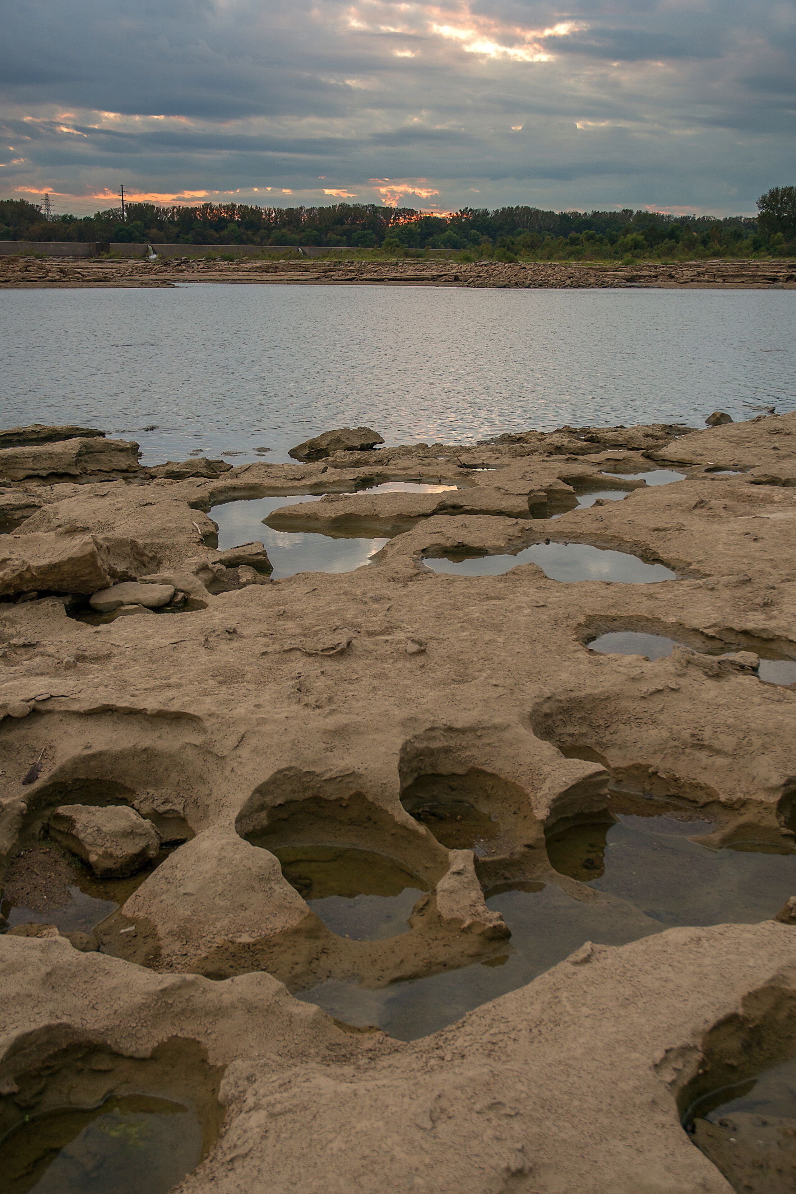 Una costa rocosa con charcos de agua y una vista distante de un atardecer sobre un área boscosa.