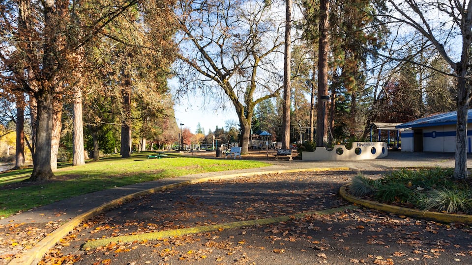 A park with a paved area, trees, a playground, and a building labeled 'Riverside Park.'