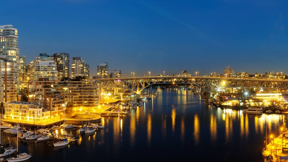 A cityscape at dusk with illuminated buildings, a bridge, and a marina with boats.