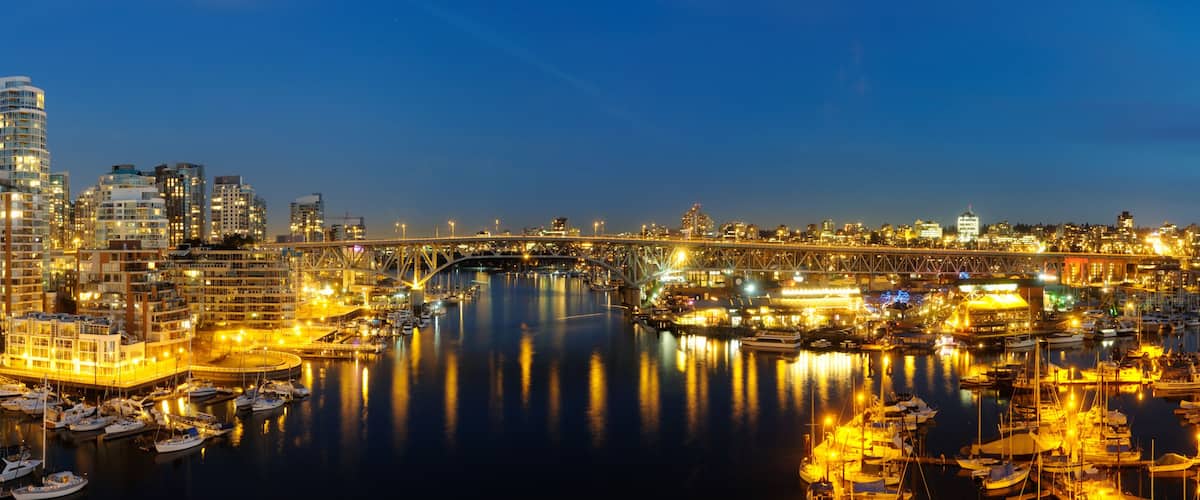 A cityscape at dusk with illuminated buildings, a bridge, and a marina with boats.