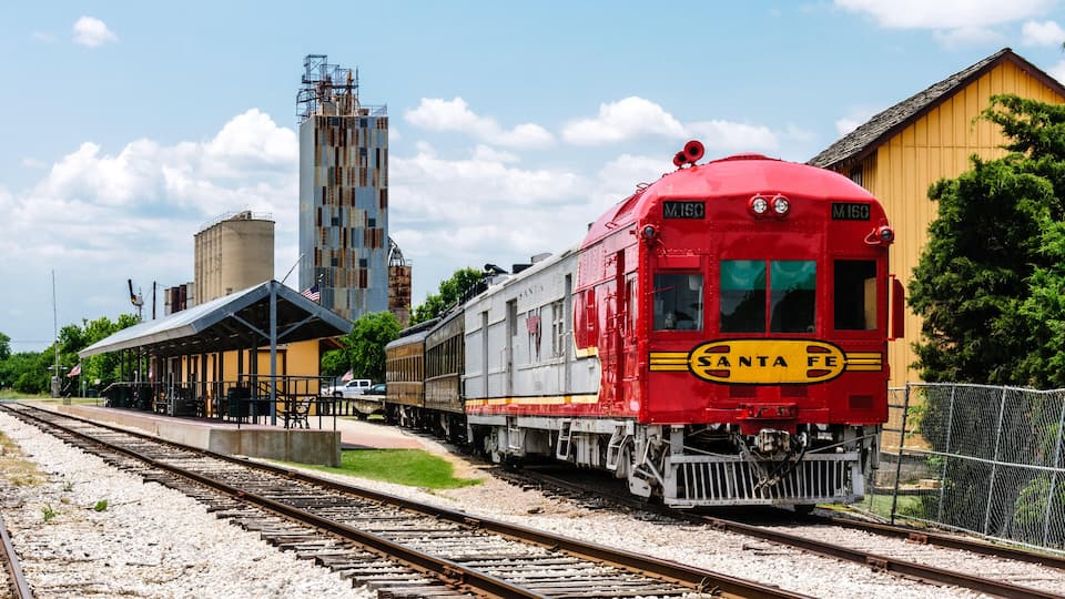 A red Santa Fe train at a station with a building and a tall structure in the background.