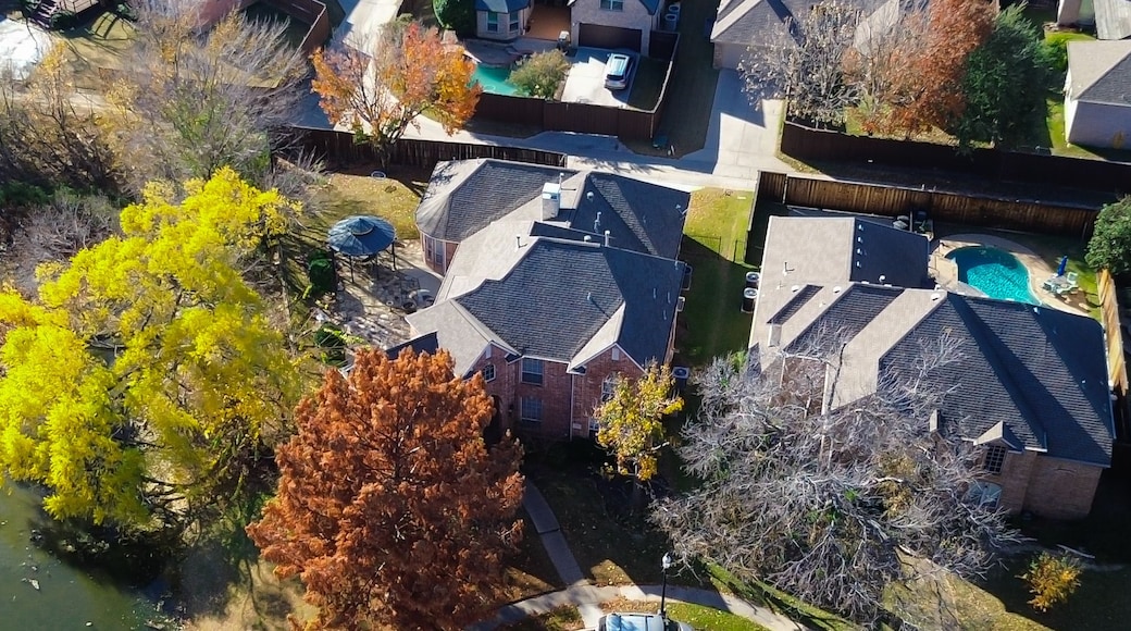 Panorama aerial view Coppell homes with pools and fenced yards line street beside lake; autumn trees in yellow and brown frame the natural shoreline and suburban edge with seasonal warmth, Texas