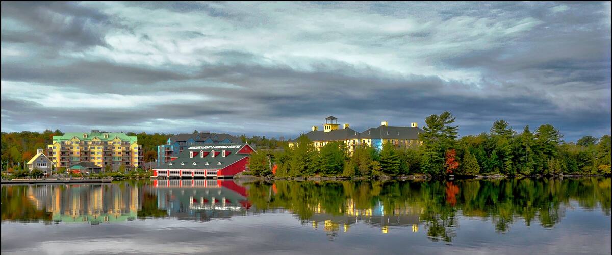 A calm lake with reflections of buildings and trees, a dock, and a red building with a green roof.