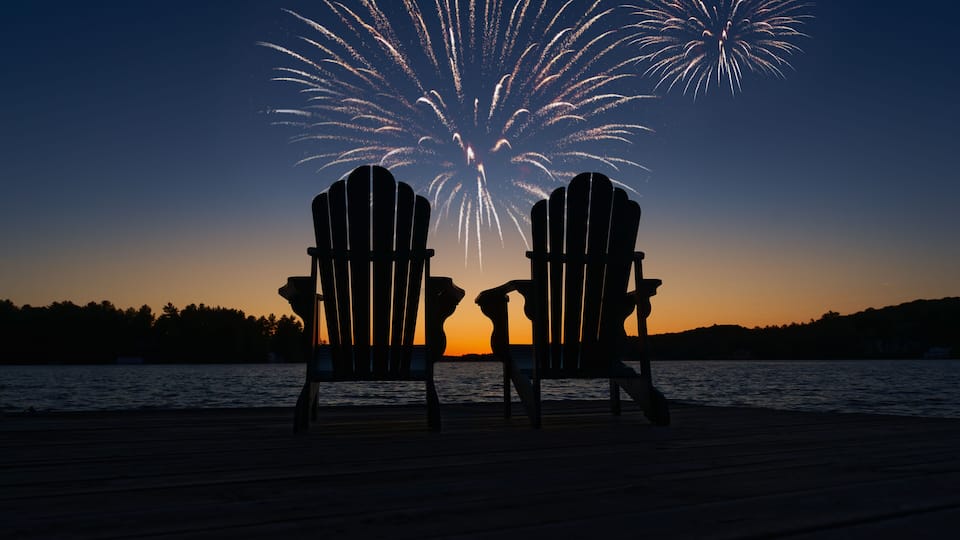Two Adirondack chairs facing a fireworks display over a body of water at sunset.
