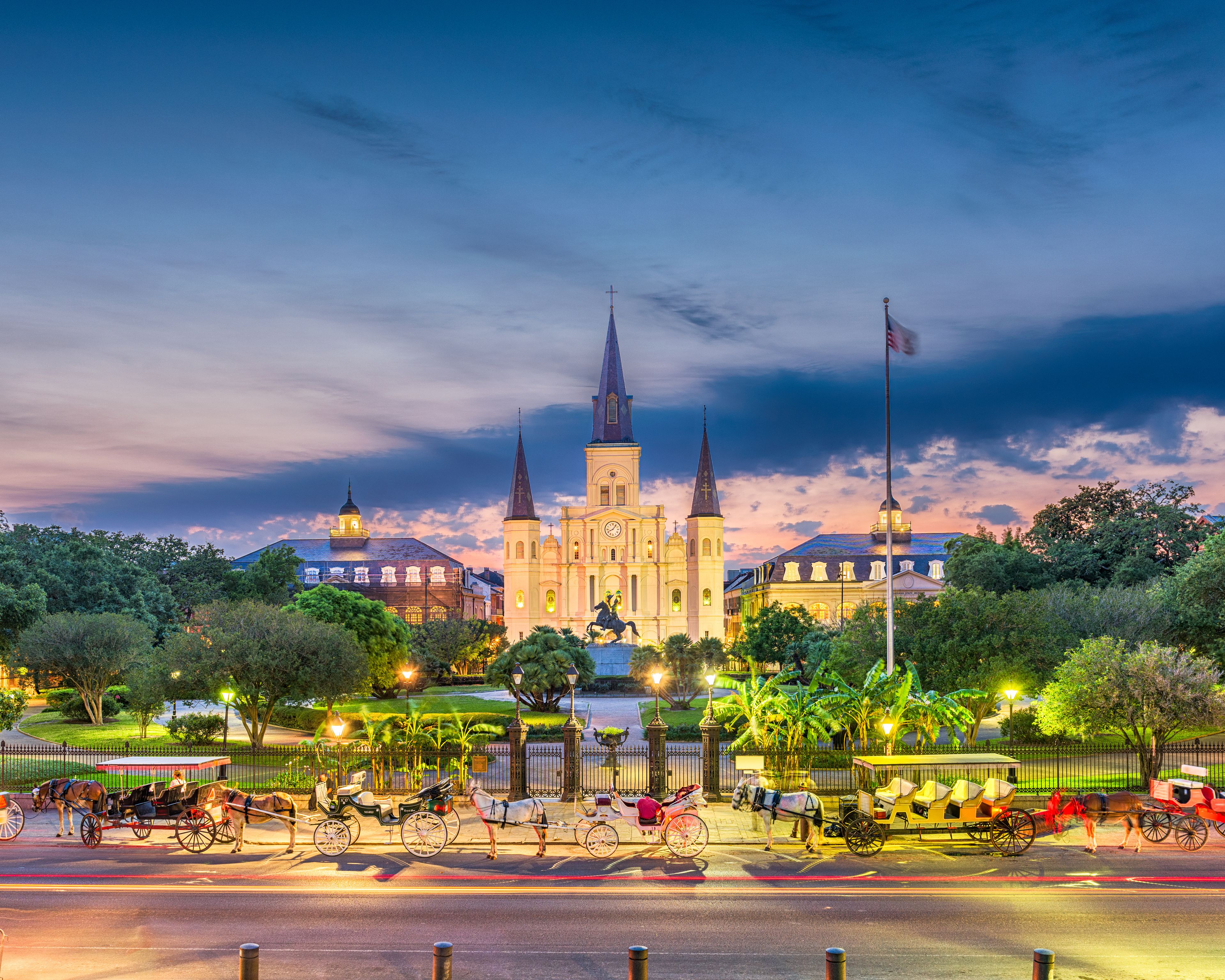 A historic church with two spires, surrounded by lush greenery and a street with horse-drawn carriages.