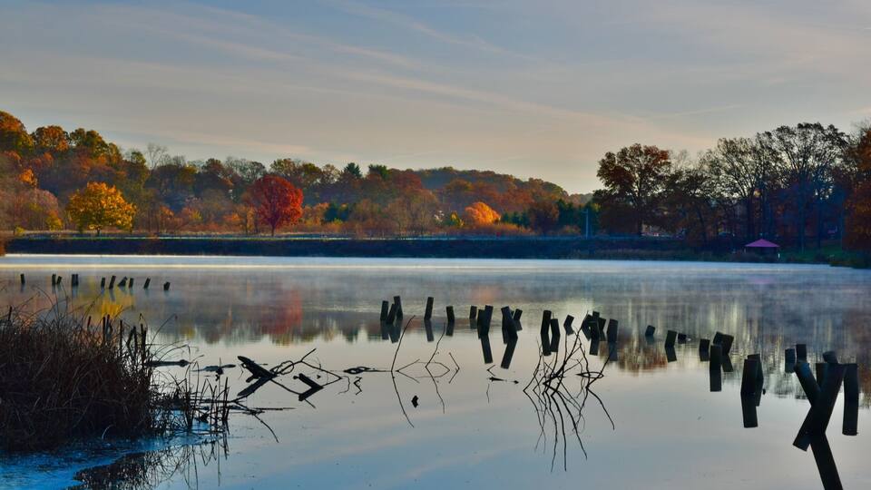 A calm lake with autumn-colored trees and wooden posts in the foreground.