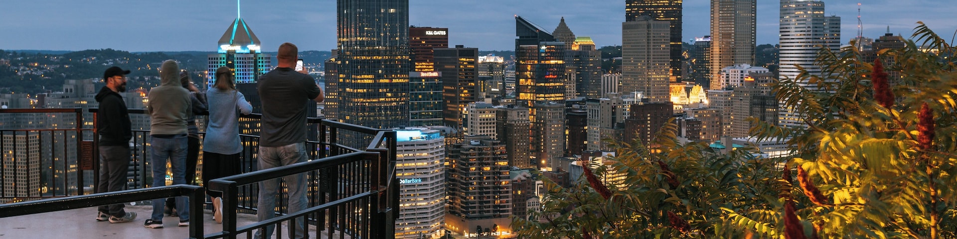 Paisagem urbana com arranha-céus modernos, incluindo um edifício em destaque com 'UPMC' e um grupo de pessoas numa varanda com vista para a cidade.