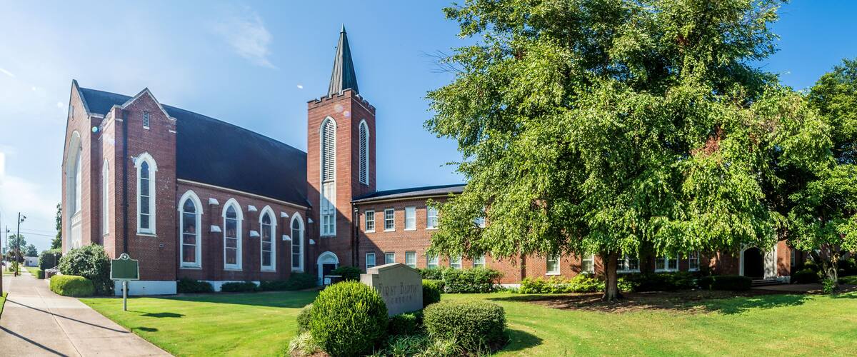 A church with a steeple and a large tree in front.
