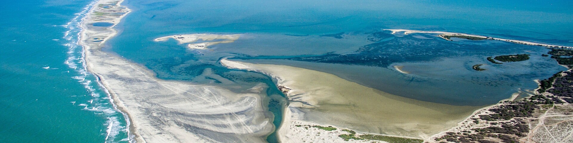 Paisagem costeira com praia arenosa, mar azul cristalino e um pequeno povoado com edifícios de telhado vermelho.