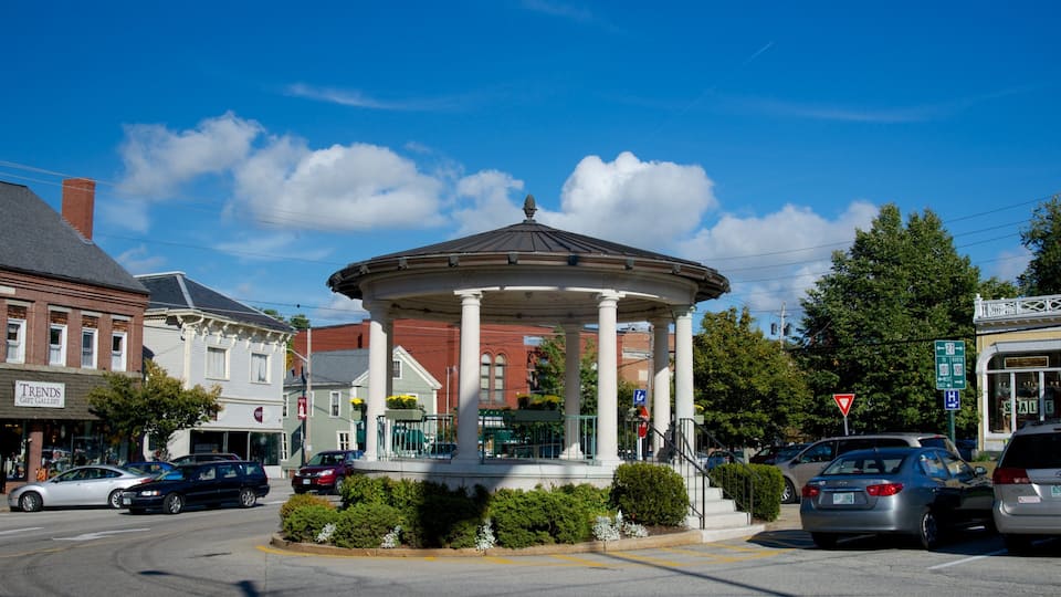 A gazebo with columns, surrounded by greenery and cars parked nearby.
